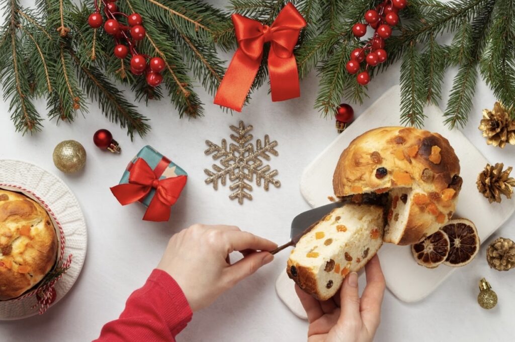 Person breaks off a piece of cake from a plate. In the background is a white table on which are coniferous branches decorated with bright red bows, around there are shiny cones