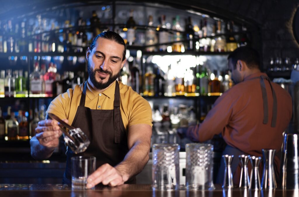 Bartender stands behind the bar and puts a piece of ice into a glass. Behind him, another bartender rearranges glasses behind the counter.