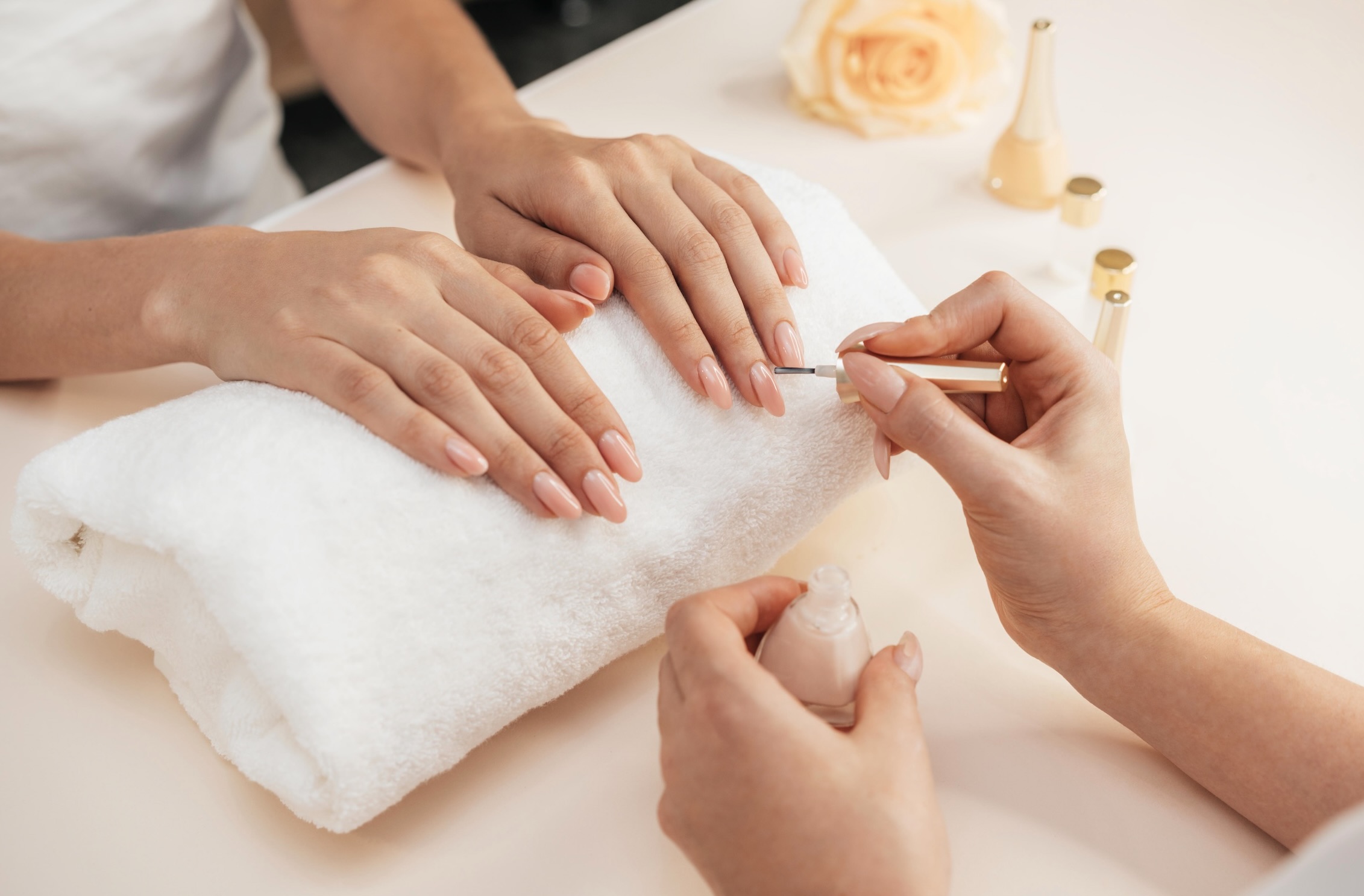 Manicurist paints the nails of a girl whose hands are resting on a towel roll. Jars of pink nail polish stand nearby.