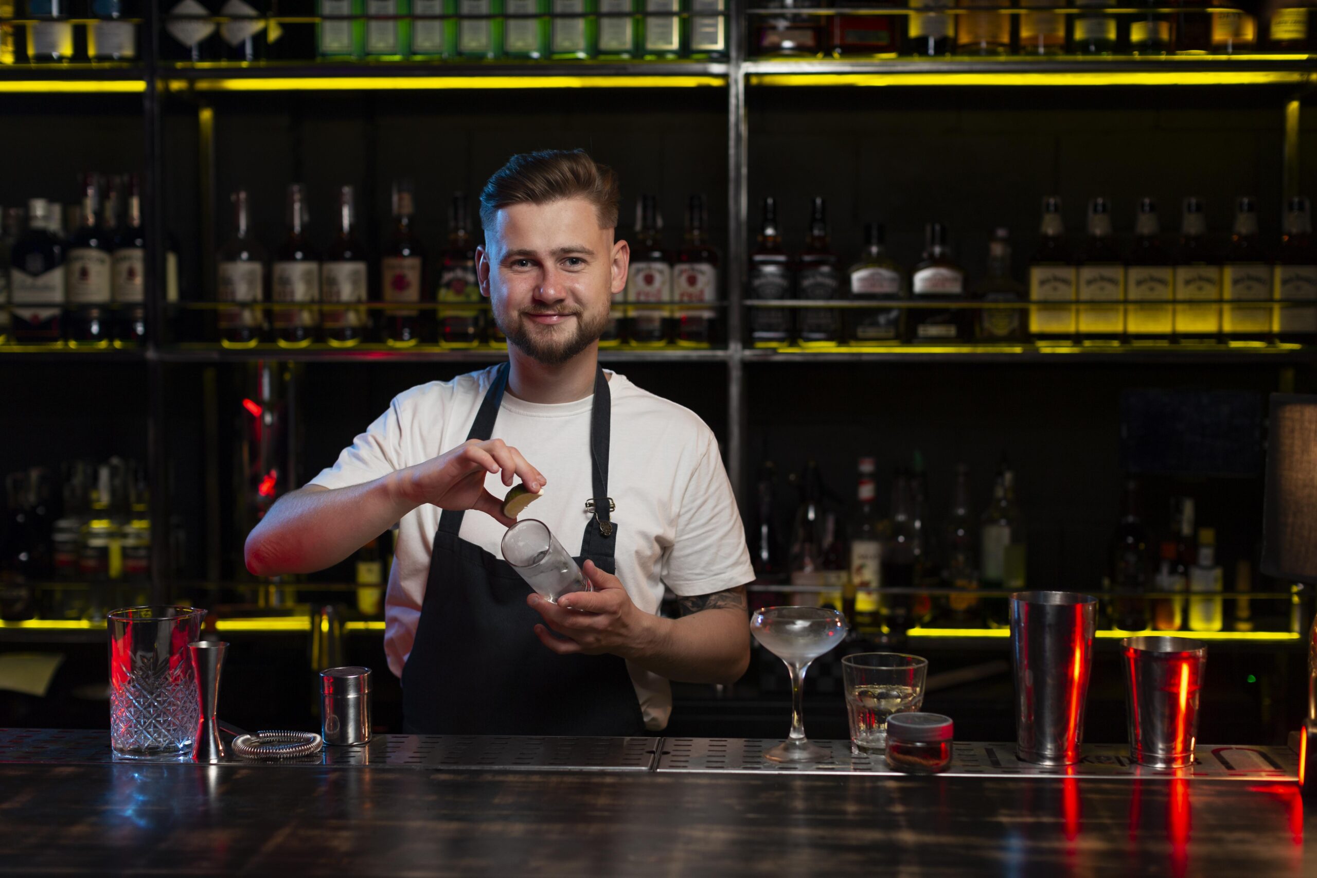 Young smiling bartender stands behind the bar counter and prepares a cocktail