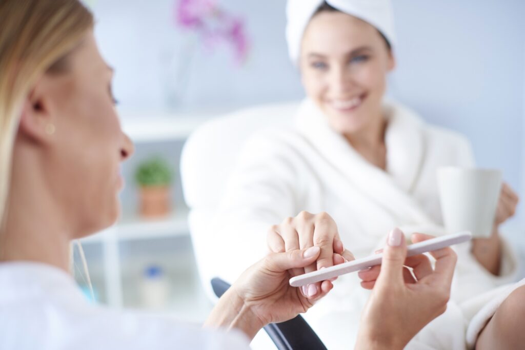 Kosmetolog wykonuje manicure – trzyma pilnik do paznokci i opracowuje dłoń klientki w białym szlafroku z ręcznikiem na głowie.