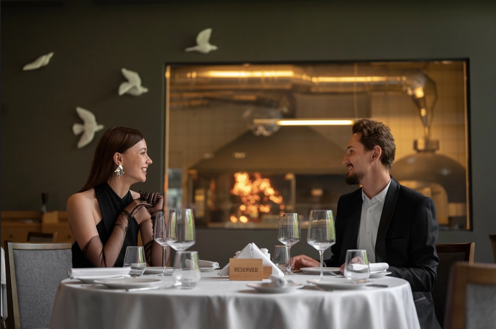 Para – kobieta w eleganckiej sukience i mężczyzna w garniturze – siedzi przy stoliku w ekskluzywnej restauracji, uśmiechając się do siebie, a na stole stoją kieliszki i talerze.