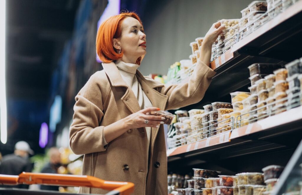 Kobieta w płaszczu stoi w supermarkecie przy półkach z suszonymi owocami i wybiera produkty.