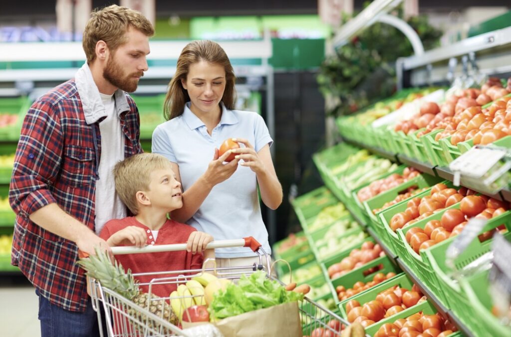 Rodzina w supermarkecie – kobieta, mężczyzna i mały chłopiec stoją przy wózku z zakupami obok półek z warzywami, kobieta trzyma pomidora.