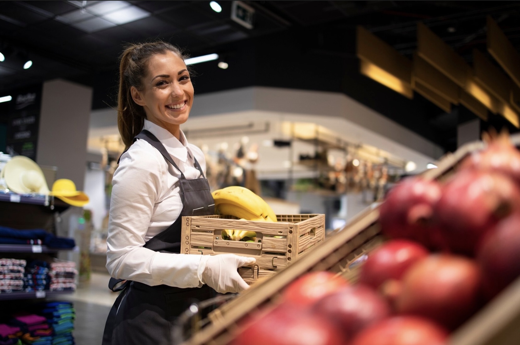 Kobieta w uniformie stoi w supermarkecie przy półkach z owocami, uśmiecha się i trzyma skrzynkę z bananami.