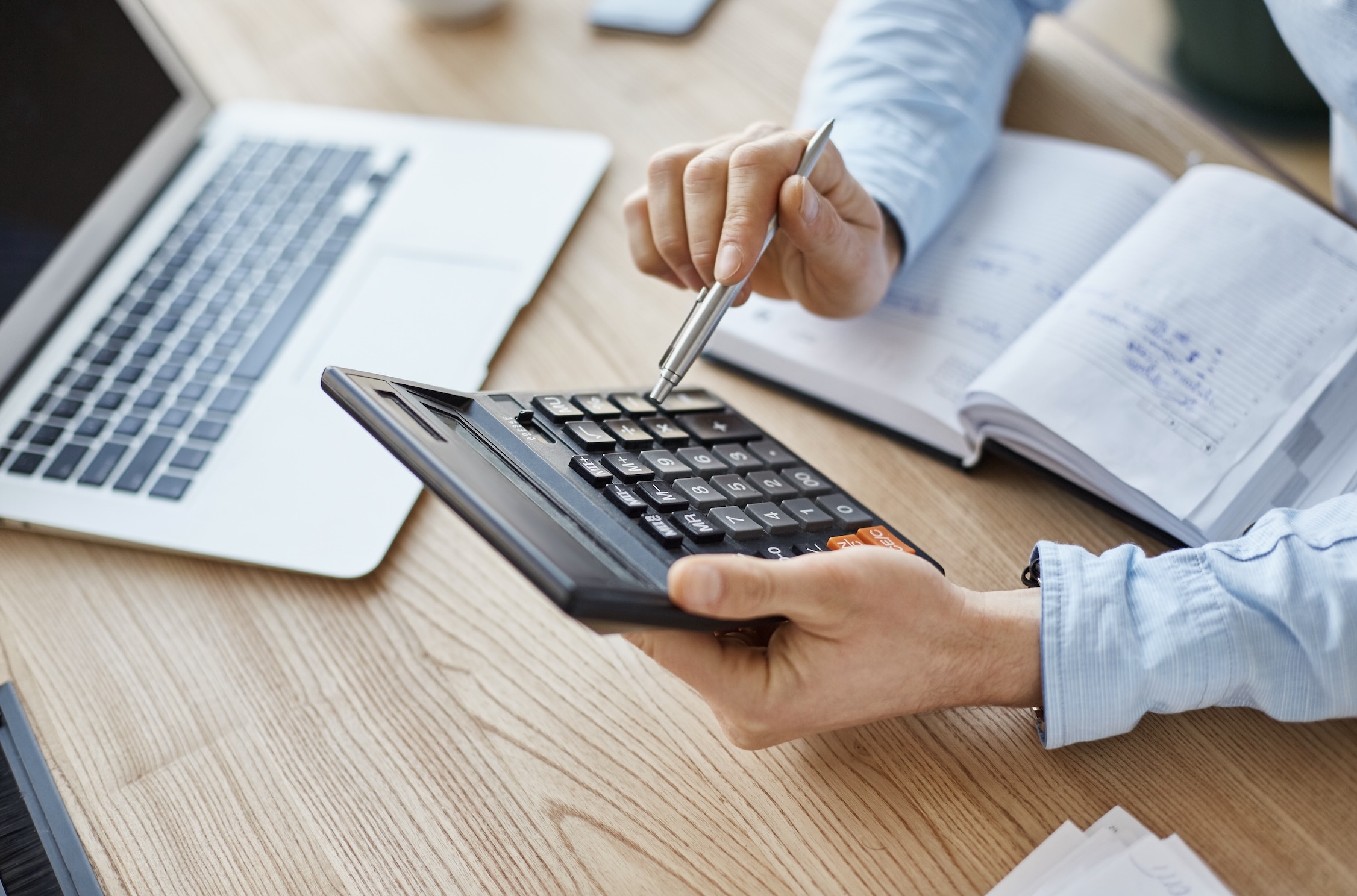 Person is holding a calculator and a pen, with a laptop and an open notebook with notes placed on the table nearby.