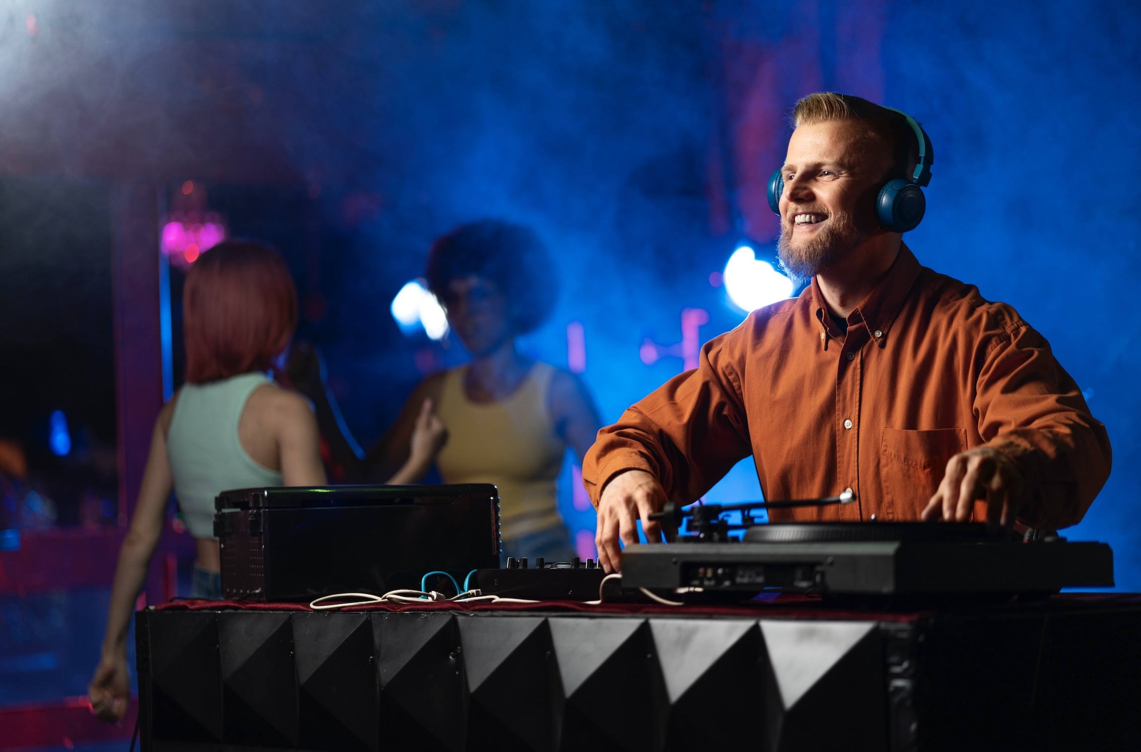 Young male DJ smiling while wearing headphones, standing behind a DJ setup, with two women dancing in the background.