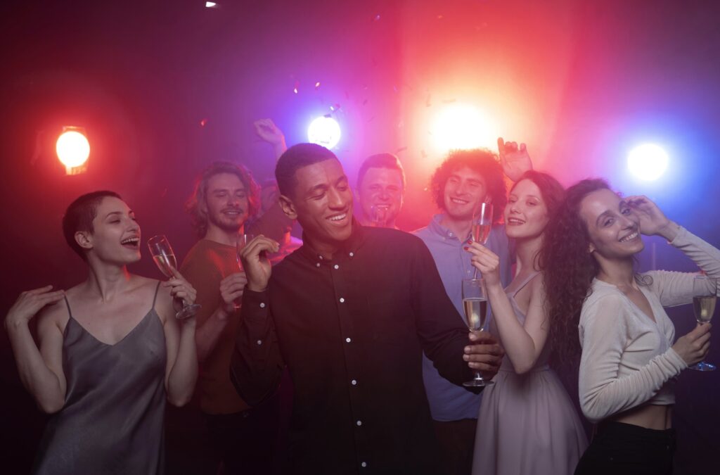 Cheerful group of four men and three women holding champagne glasses, dancing in the dark under red and blue spotlights.