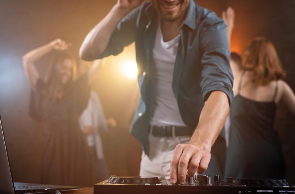 Smiling man in a shirt and white pants stands behind a DJ setup, with women in dresses dancing in the background.