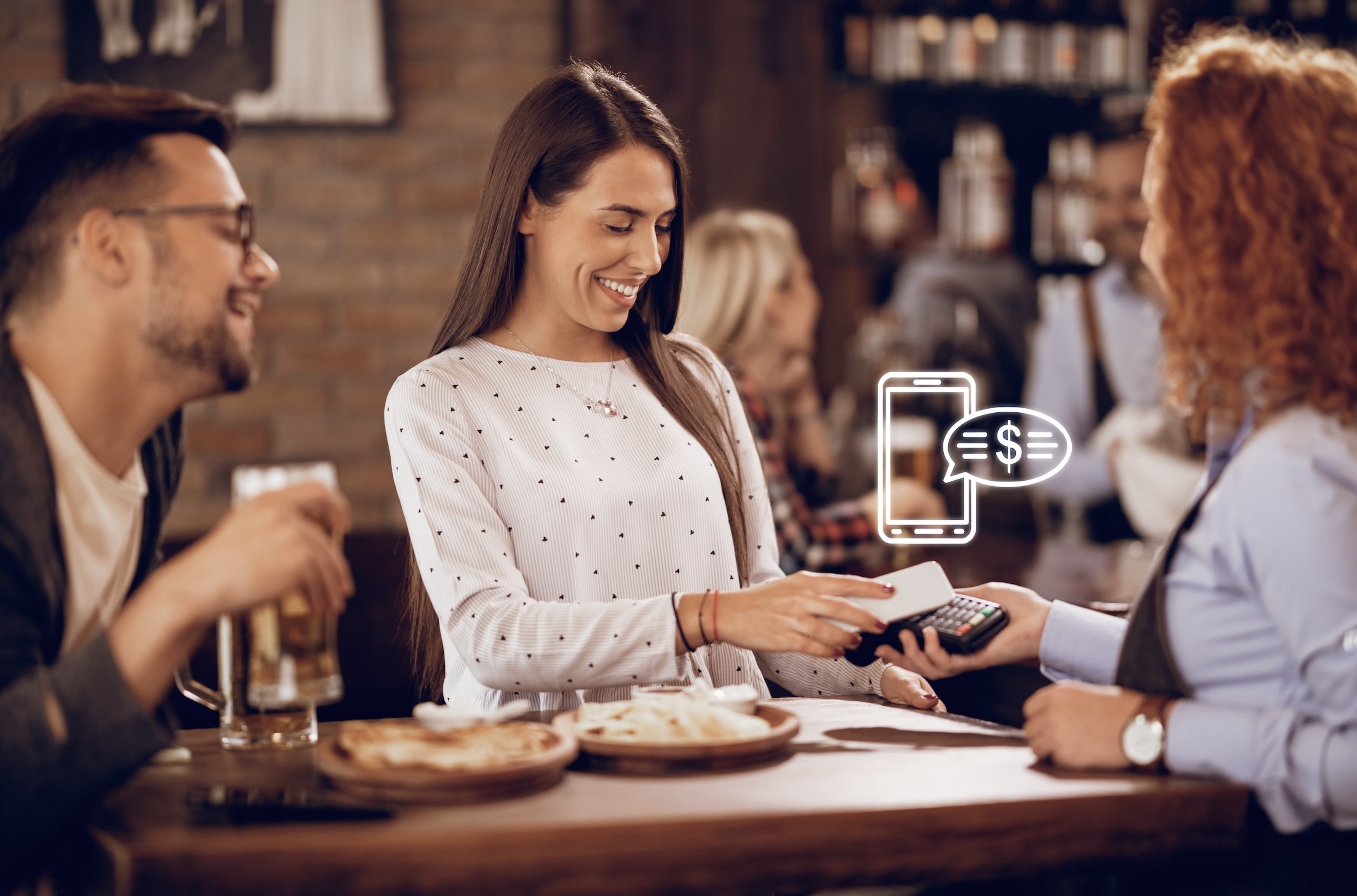 A young woman is smiling while paying the bill with her smartphone using a contactless terminal held by a waitress in a bar, as a man with a beer sits next to her.