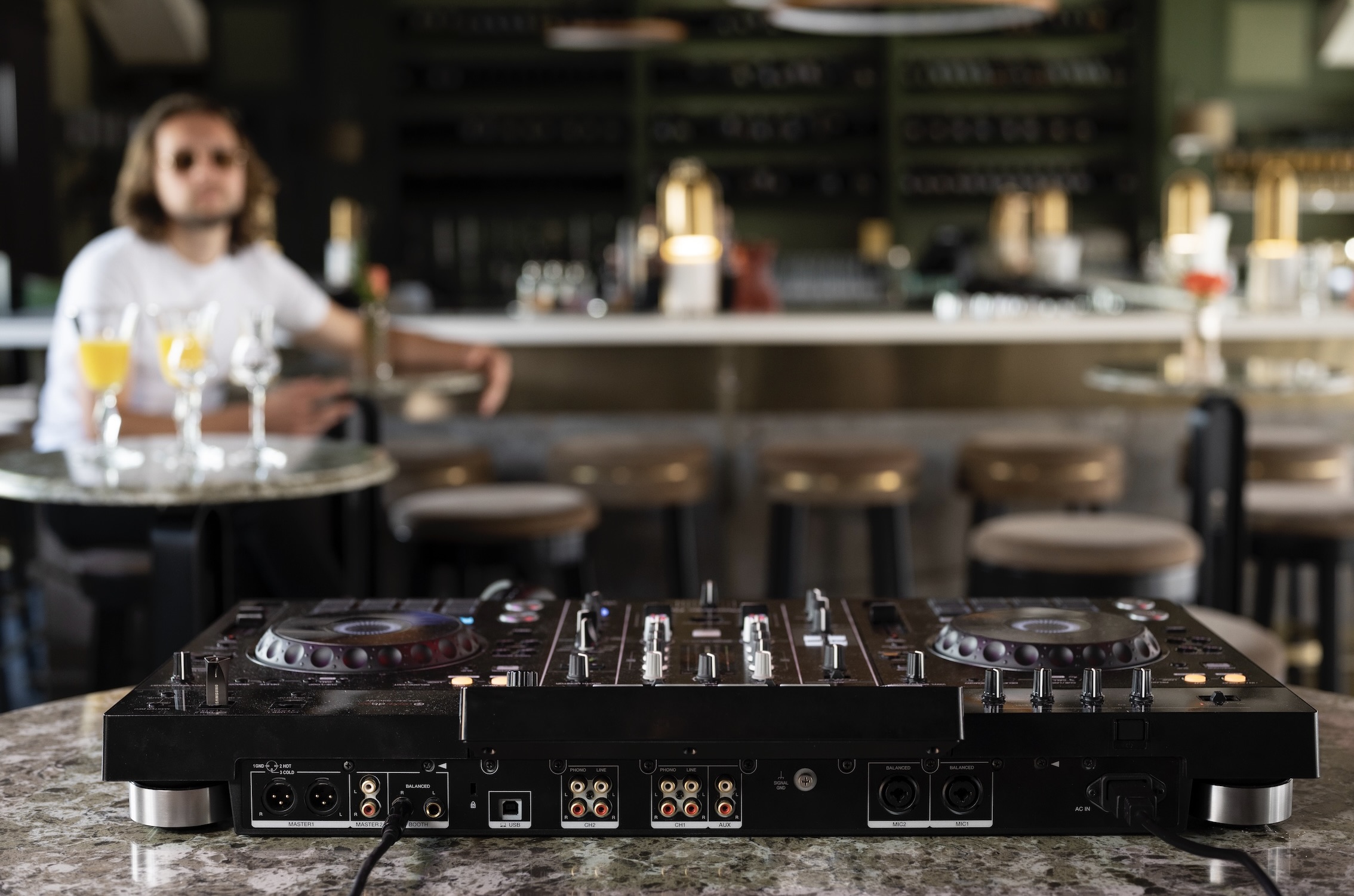 Professional DJ mixer is placed on a marble table in a modern bar with blurred stools, glasses of orange drinks, and a guest in the background.