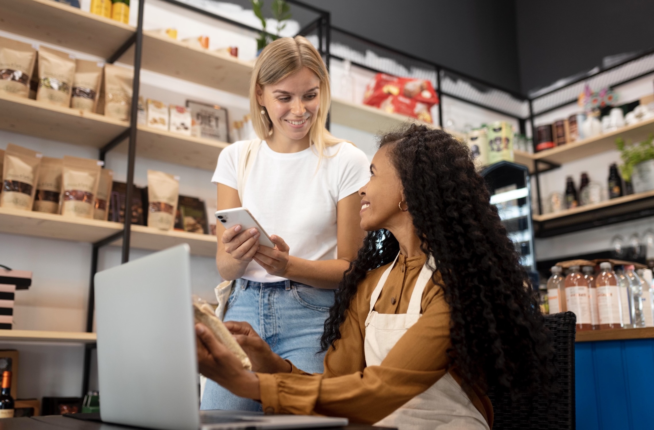 Two young women, a blonde and a brunette, smiling and talking; brunette sits at a desk with a laptop, blonde stands holding a smartphone, store shelves in the background.