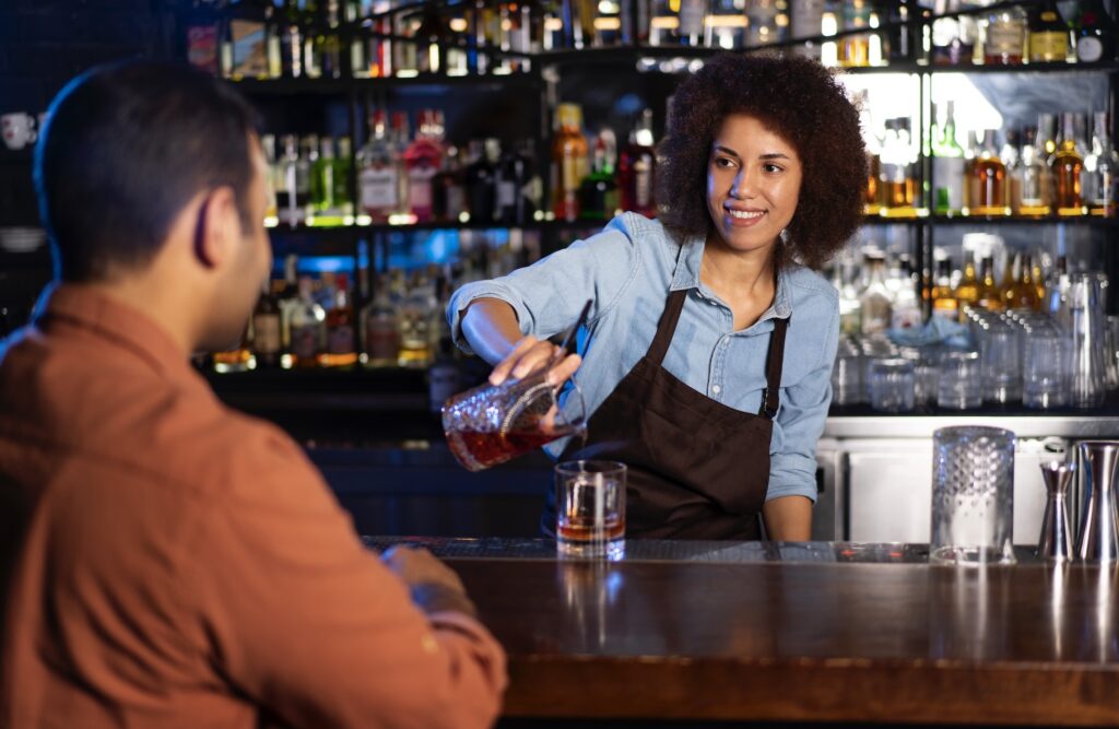 Female bartender in an apron pouring a cocktail behind the bar, shelves with bottles behind her, man in a shirt sitting at the bar.