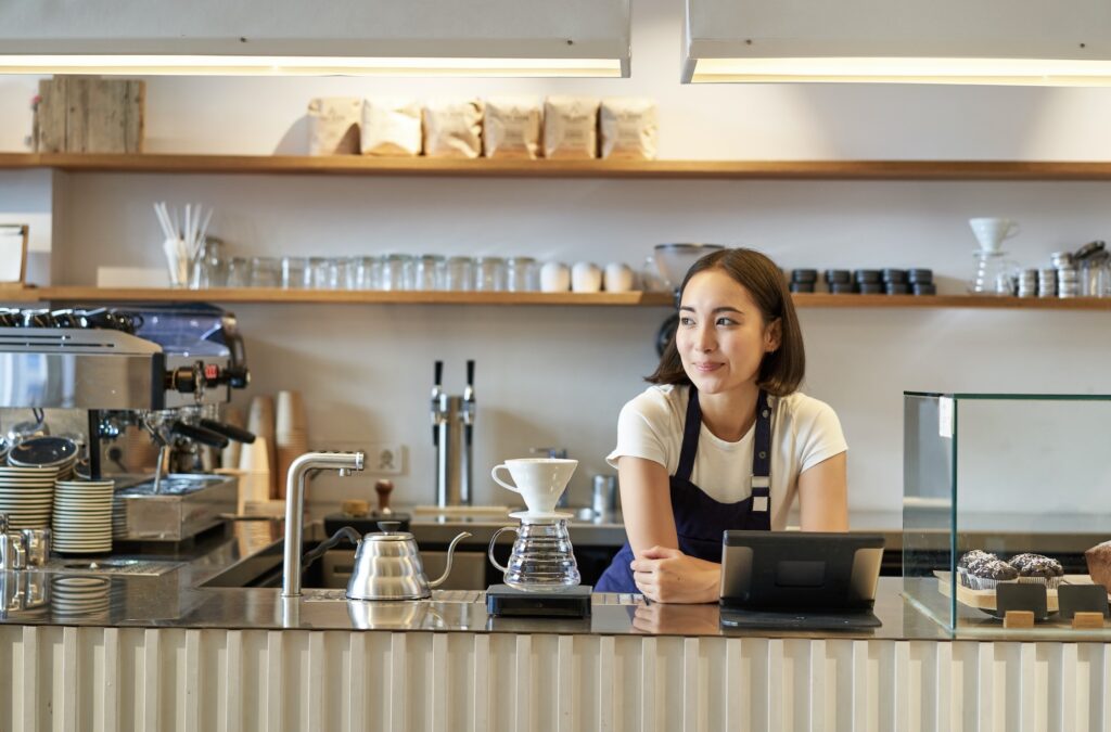 Smiling female barista in an apron behind a café counter, with a coffee machine, filters, pastry shelves, and café equipment in the background.