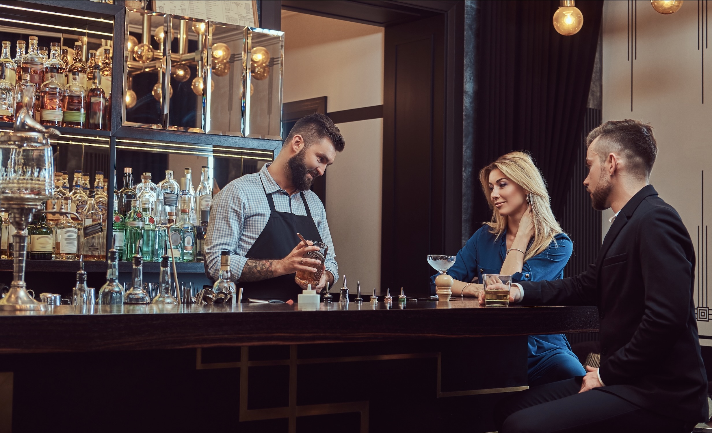 Bartender preparing a cocktail behind the bar while a man and a woman sit across the counter enjoying their drinks.