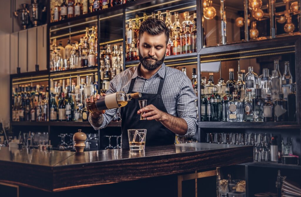 Bearded bartender in an apron preparing a cocktail behind a bar lined with bottles.