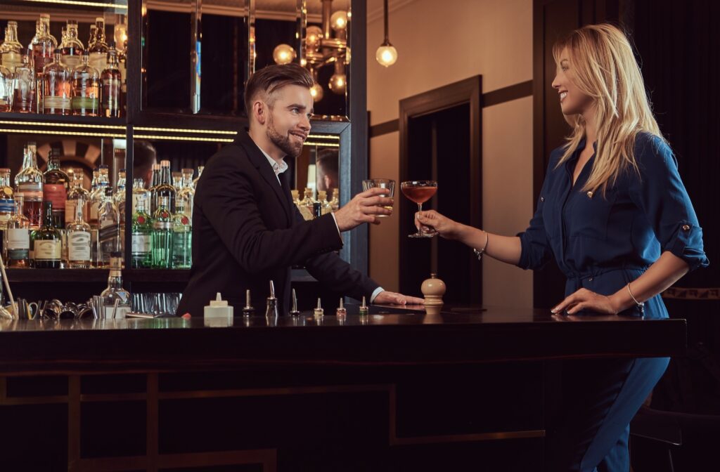 Man standing behind a bar holding a drink and clinking glasses with a blonde woman holding a cocktail on the other side of the counter.