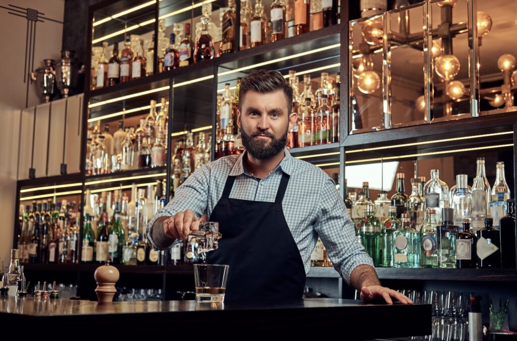 Bearded bartender in an apron pouring liquid into a glass behind a bar lined with bottles.