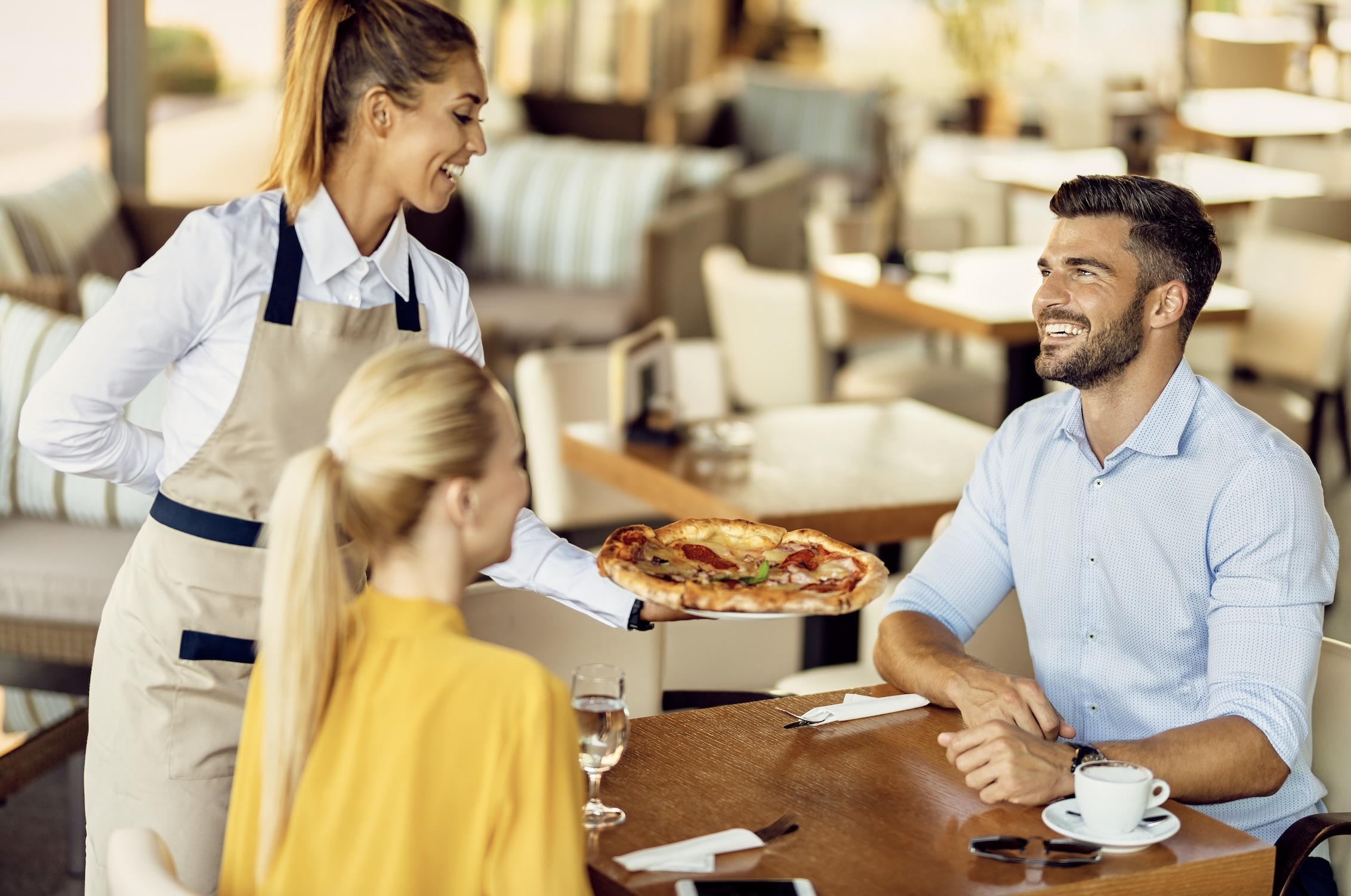 Smiling man and woman sitting at a restaurant table with a glass and a cup, while a waitress places a pizza dish on the table.