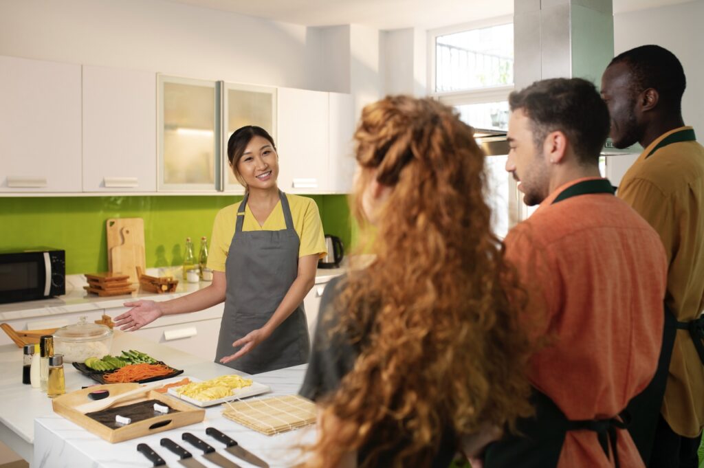 Smiling woman in an apron standing in a kitchen, pointing with both hands to a table with kitchen utensils, spices, plates, and cutting boards with chopped vegetables, while two men and a woman in waiter uniforms stand in front of her with their backs to the camera.