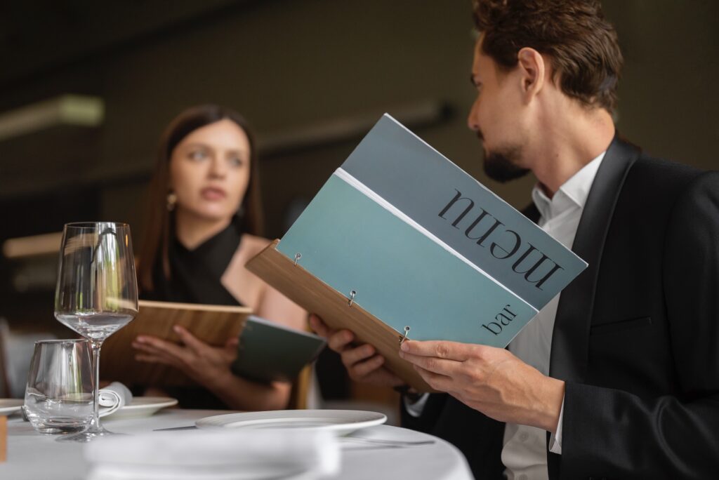 Man and woman sitting at a restaurant table holding menus, with glasses and plates on the table.