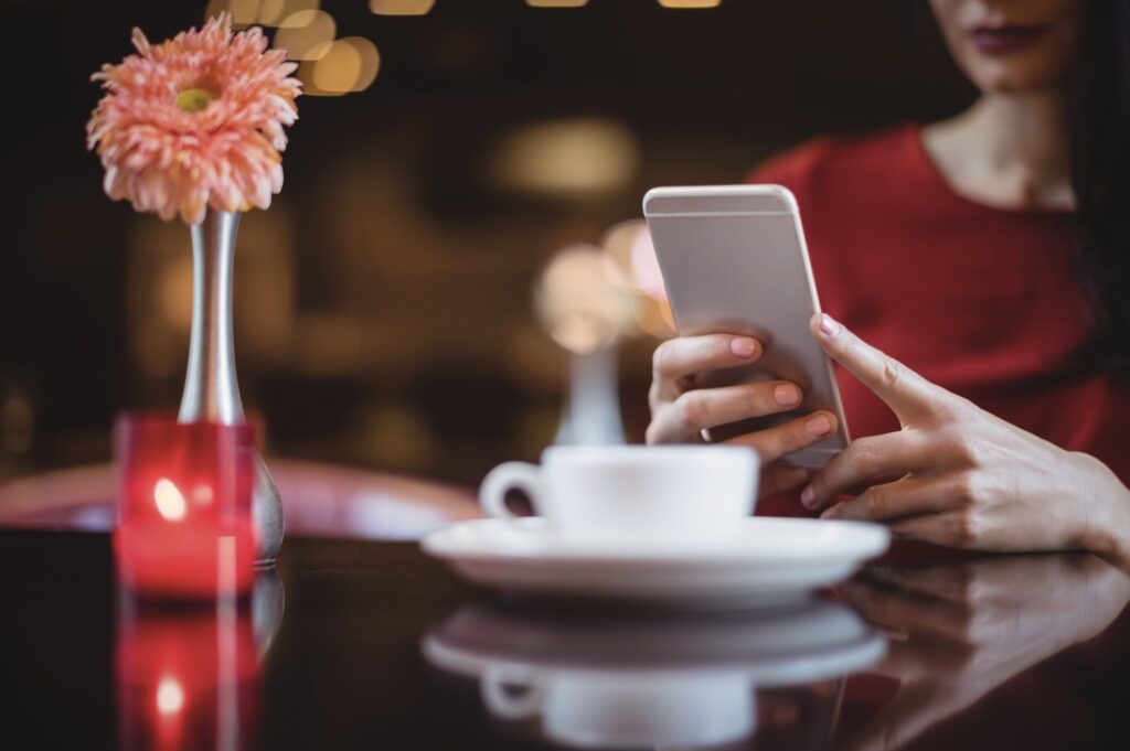 Close-up of a woman sitting at a table, showing only her lower face and hands holding a phone, with a white coffee cup and a chrysanthemum flower in a vase in the foreground.