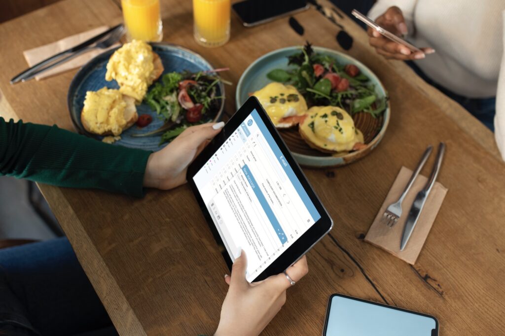 Restaurant table with two plates of scrambled eggs with vegetables, two glasses of orange juice, and two sets of cutlery, with a person on the left holding a tablet with a report and another on the right holding a phone.