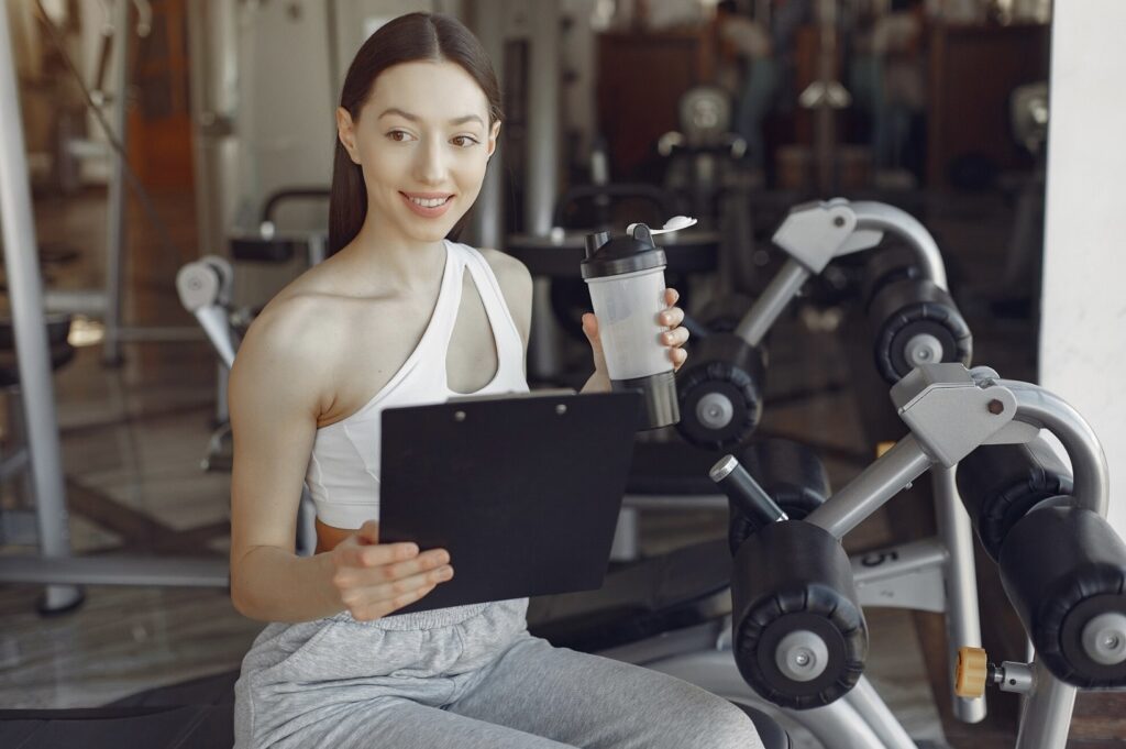 Woman in a light tracksuit sitting on a gym machine, holding a thermal mug and a black tablet in her hands.