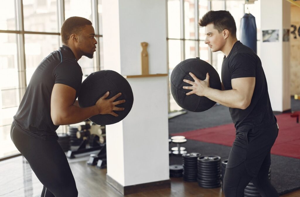 Two men in black sportswear standing opposite each other in a gym, slightly bent over and holding large black balls.