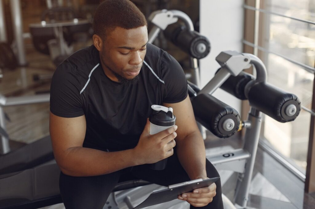 Man in a black tracksuit sitting on a gym machine, holding a thermal mug and a black tablet in his hands.