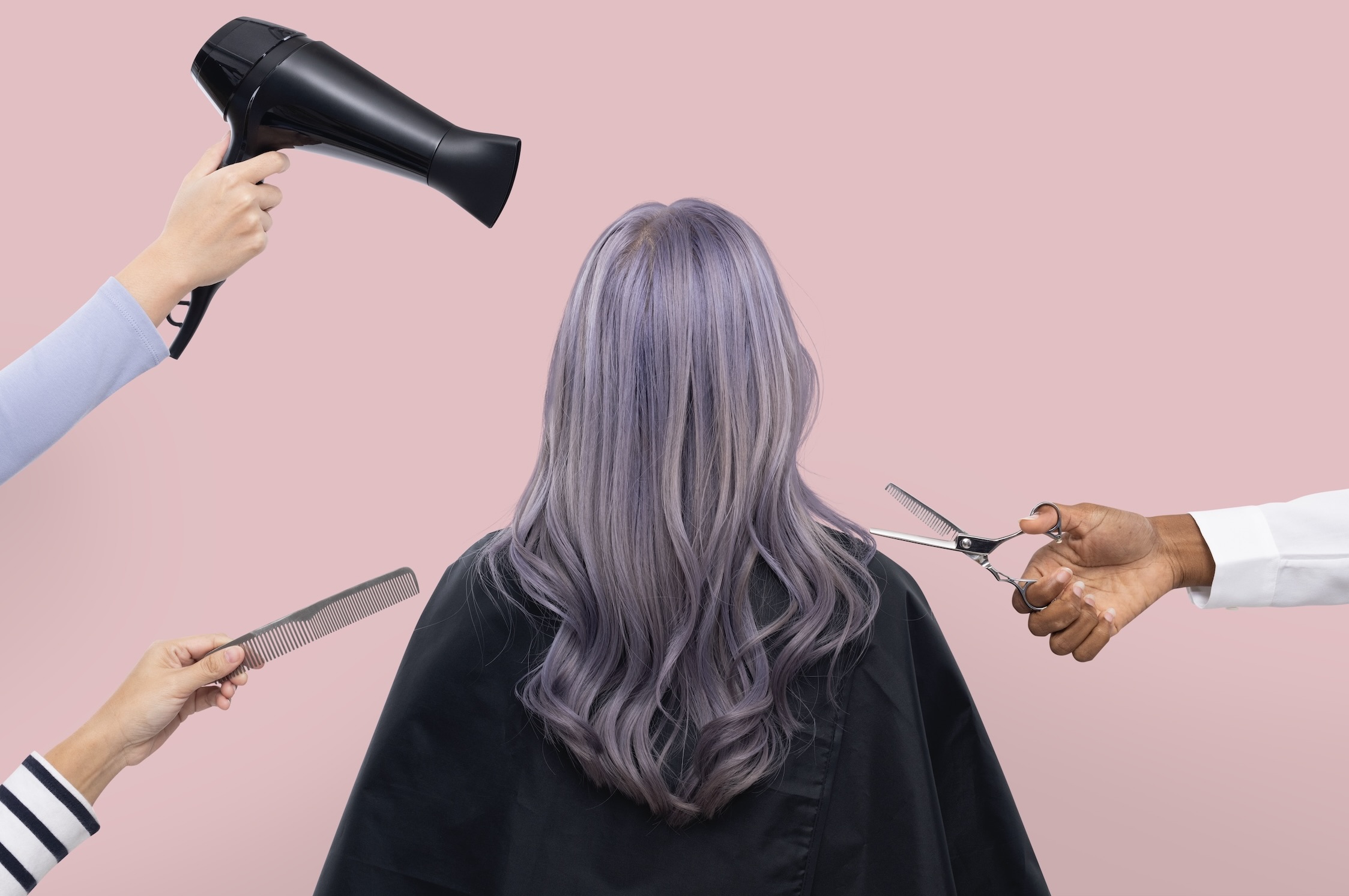 On a pink background, a woman with lavender hair and a black cape sits with her back to the camera, while hands holding a hairdryer, a comb, and scissors reach toward her from different sides.