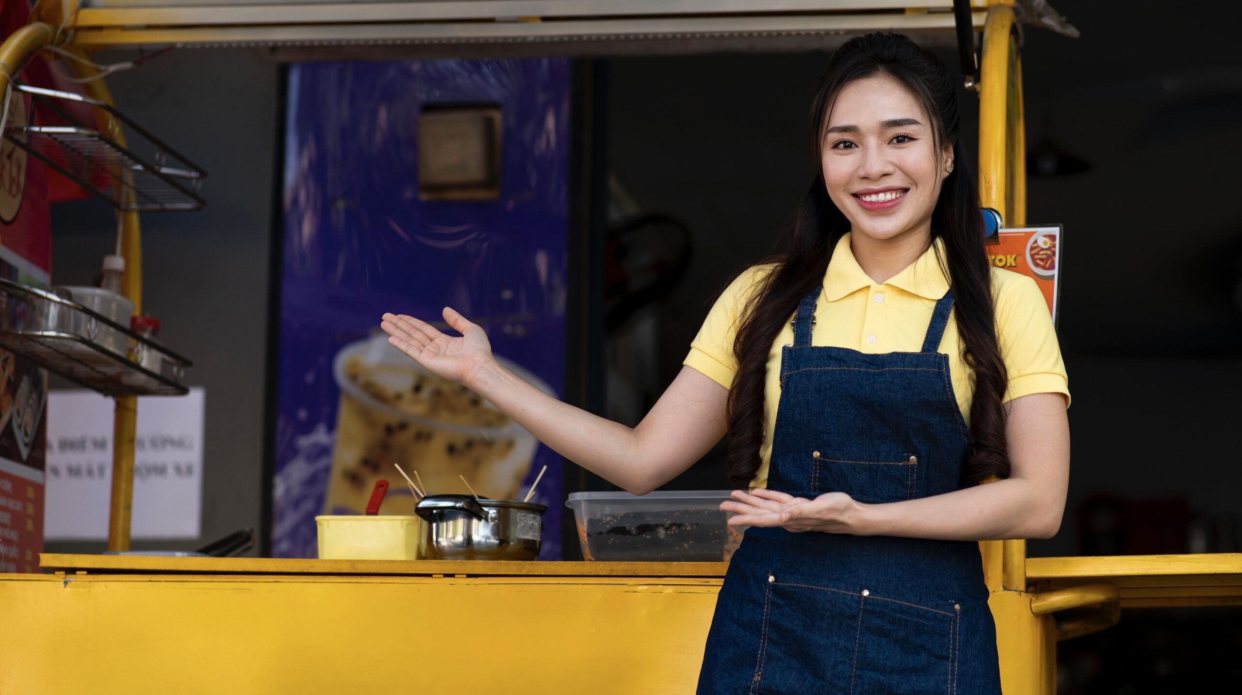 Young woman in an apron smiling and pointing at a food truck, which is visible in the background.