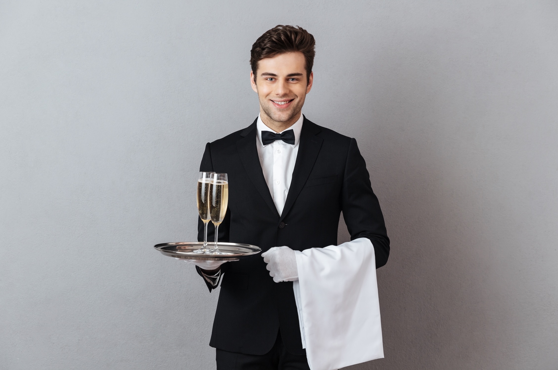 Young man in a waiter uniform standing against a gray background, smiling and holding a tray with two champagne glasses in one hand and a white towel draped over the other arm.