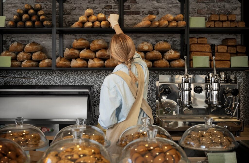 A bakery worker reaches for artisan bread on wooden shelves behind a pastry counter filled with fresh baked goods.