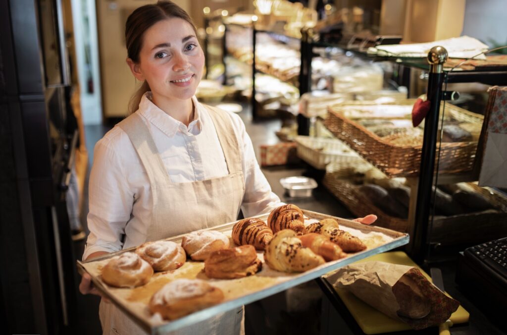 A bakery staff member presents a tray of assorted pastries, including croissants and buns, inside a cozy bakery interior.