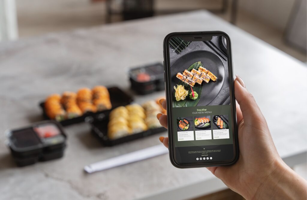 A person holding a smartphone displaying a sushi menu app with takeaway sushi trays blurred in the background on a kitchen counter.