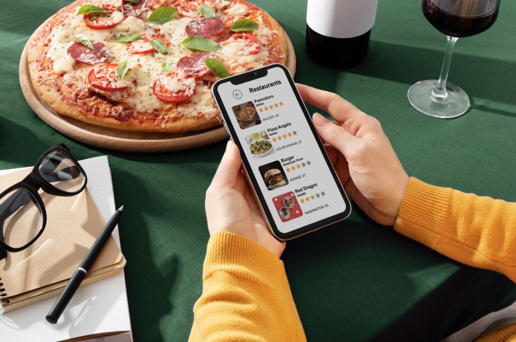 A person using a restaurant finder app on a smartphone while sitting at a table with a large pizza, a glass of red wine, and a notebook.