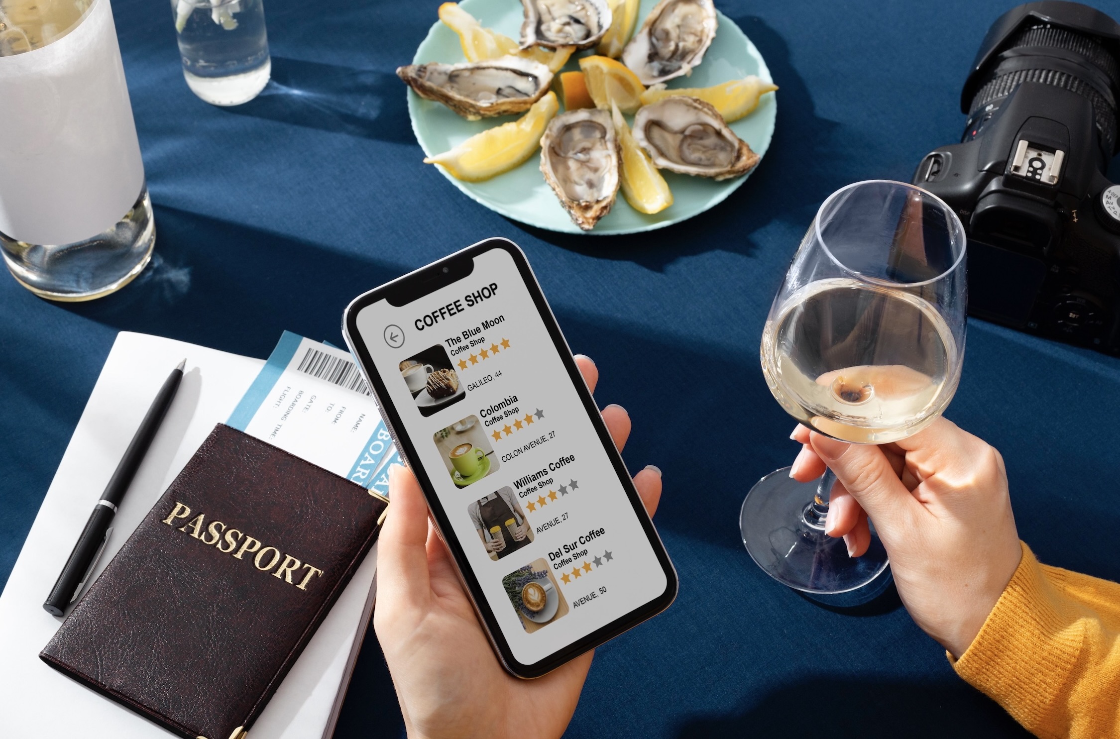 A traveler holding a smartphone with a coffee shop app open, sitting at a table with oysters, a glass of white wine, a passport, and a camera.