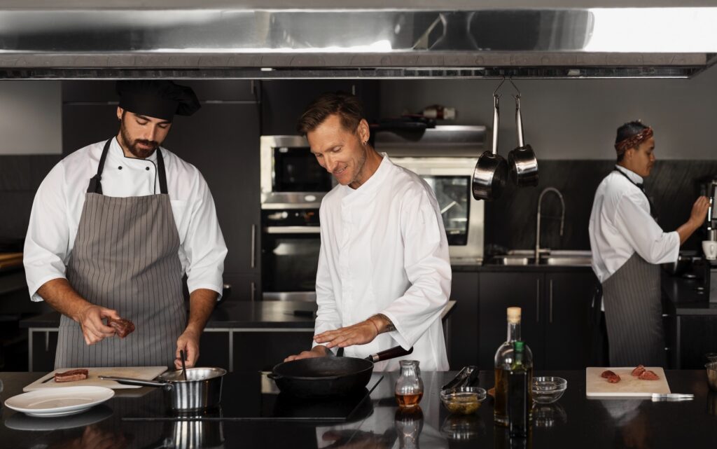 Three male chefs wearing white coats and aprons work at different stations in a professional stainless steel kitchen.