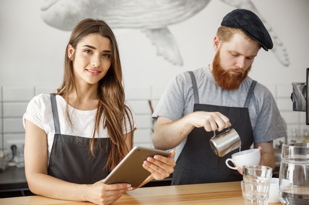 A smiling female barista holds a tablet while a male barista with a red beard pours latte art into a cup in a cafe setting.