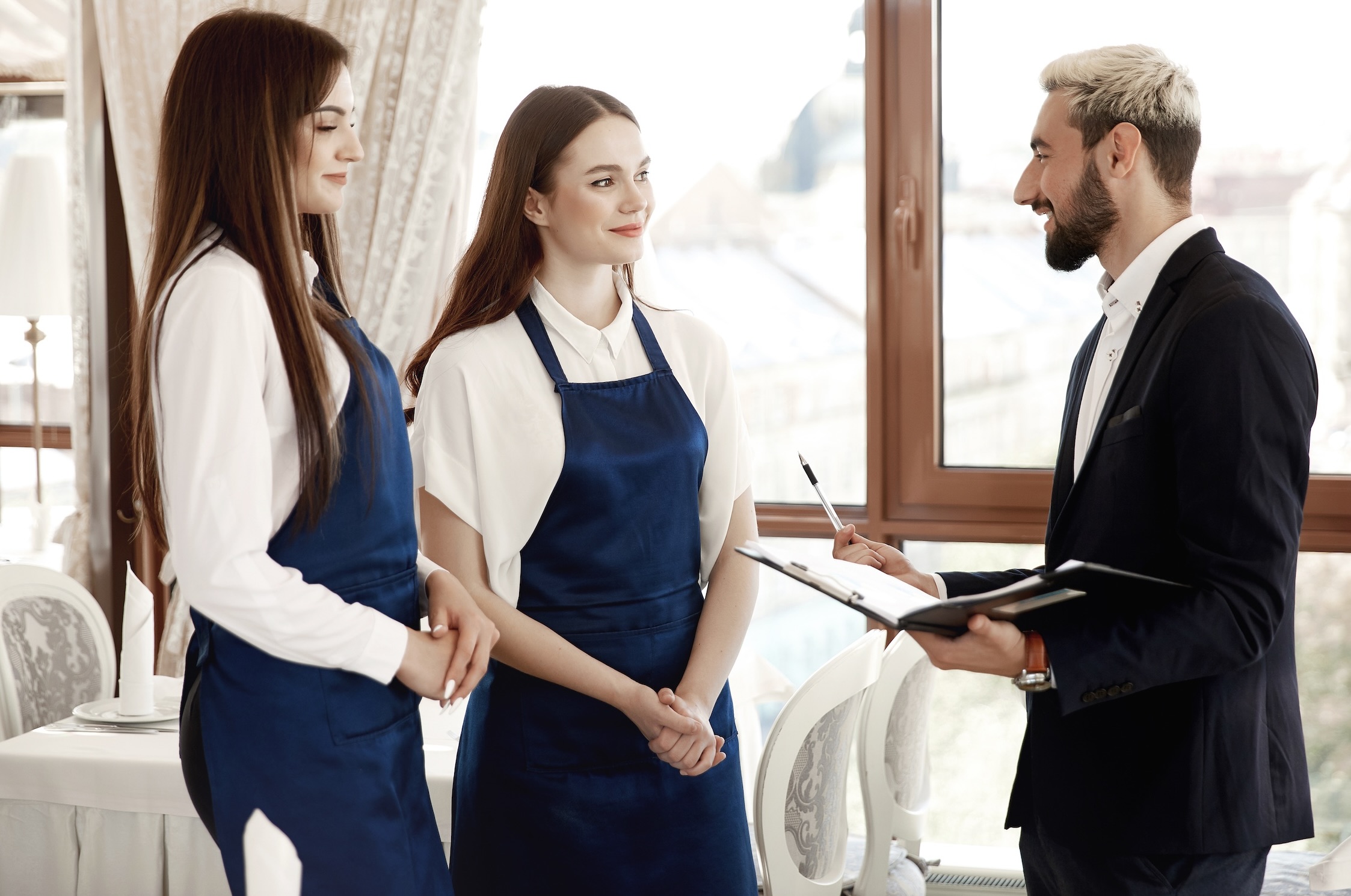 A male restaurant manager holding a clipboard speaks to two female waitresses wearing blue aprons.