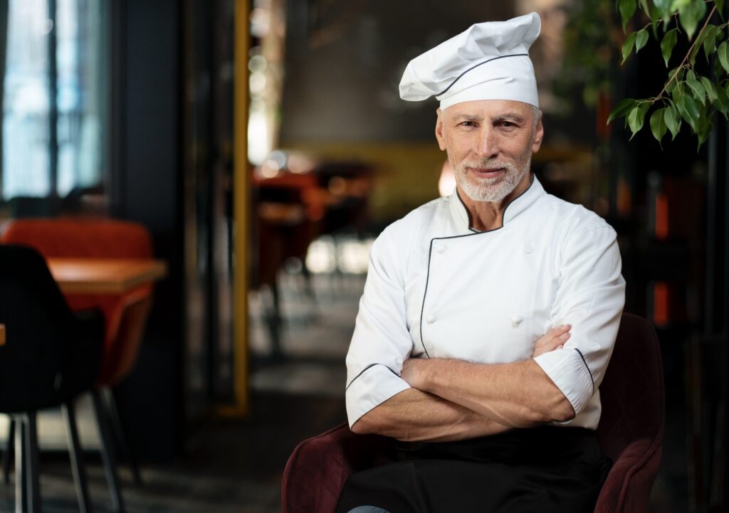 A confident, gray-haired male chef in a white uniform and hat sits with his arms crossed inside a modern restaurant.