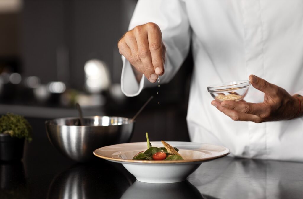 A chef in a white uniform sprinkles seasoning over a small, elegant dish in a commercial kitchen setting.