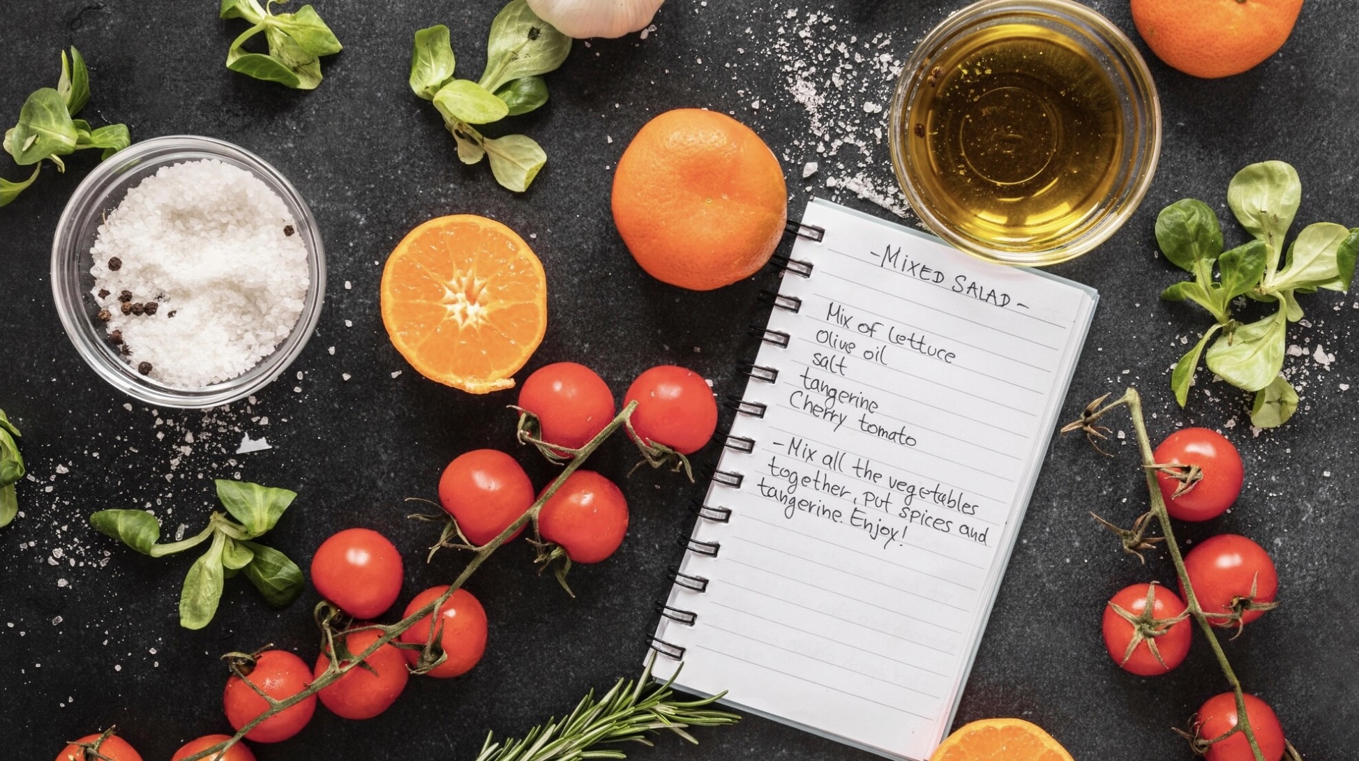 Ingredients for a mixed salad including cherry tomatoes, tangerines, and a handwritten recipe on a black surface.