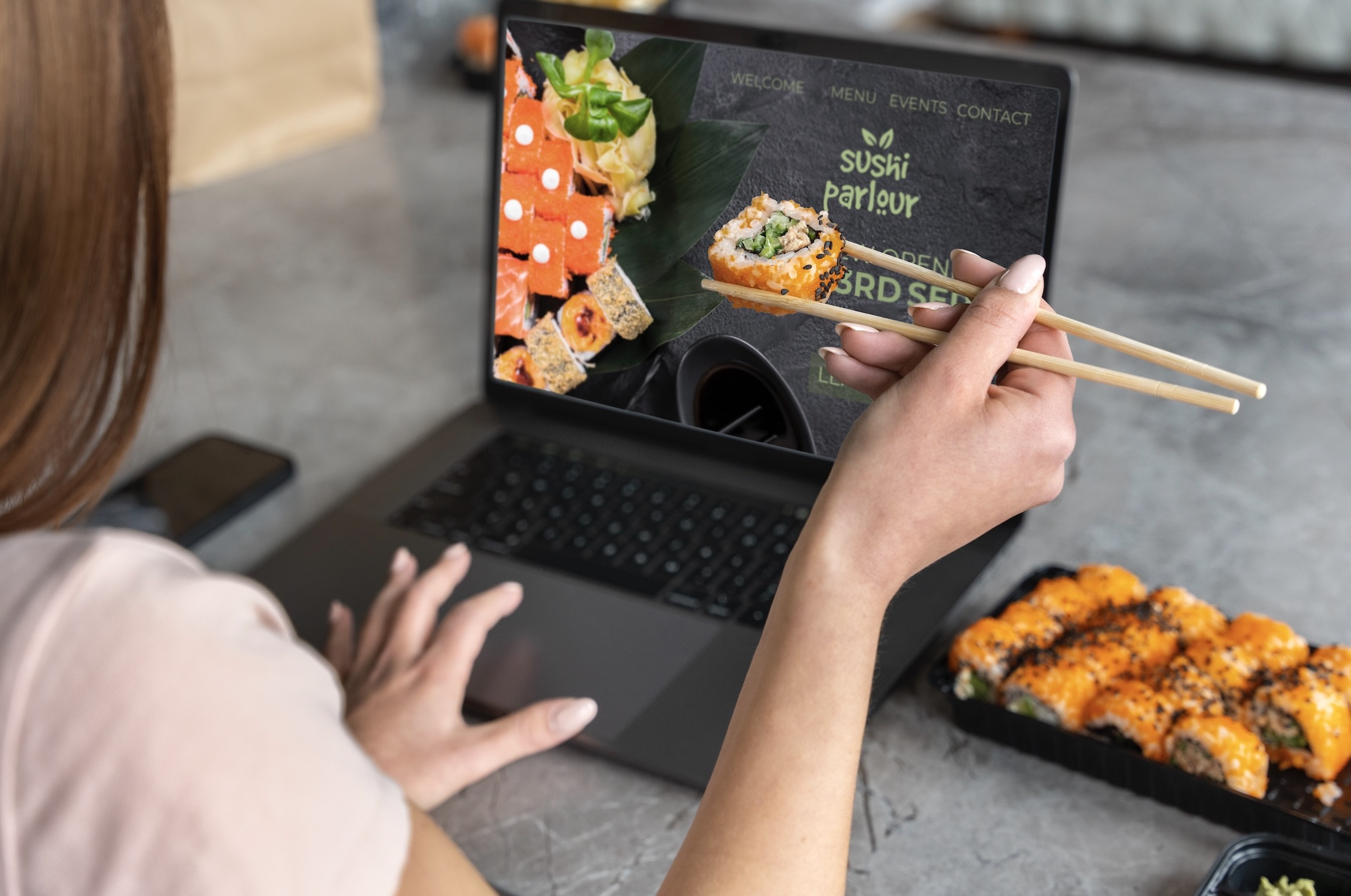 A person uses chopsticks to hold a piece of sushi while looking at a sushi restaurant's website on a laptop.