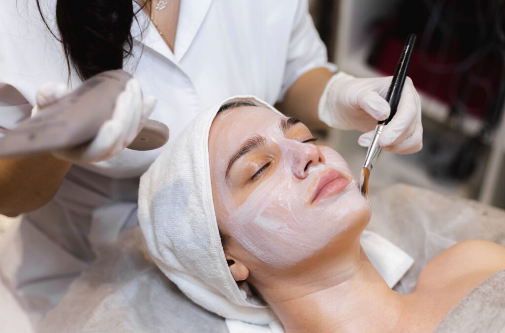 A woman receives a white facial mask application from a beautician using a brush in a spa setting.