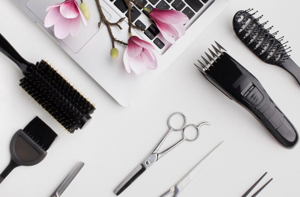 Various black hair styling tools including brushes, clippers, and scissors are arranged with pink flowers on a white surface next to a laptop.