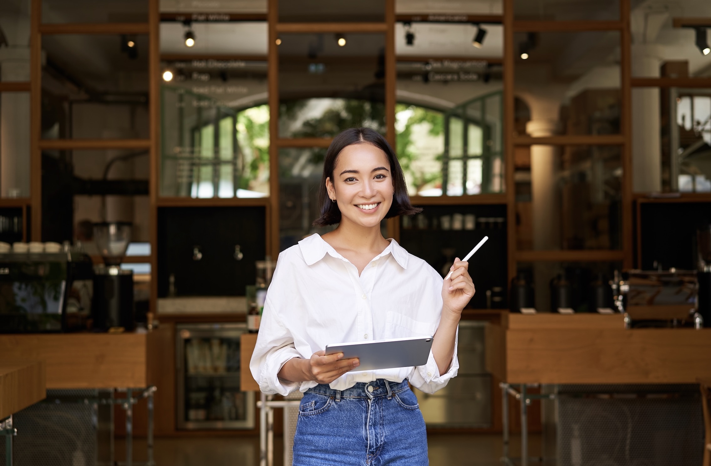 Smiling young woman with short hair stands in a modern cafe wearing a white shirt and jeans, holding a tablet and stylus.