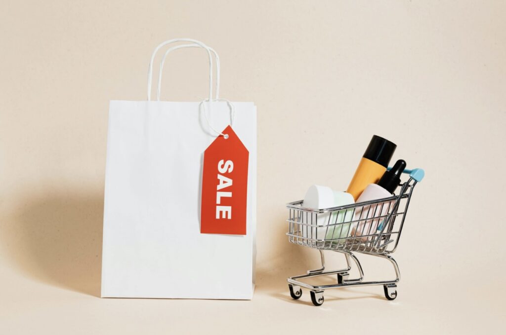 A white paper shopping bag with a red SALE tag stands next to a small metal shopping cart filled with cosmetics against a beige background.