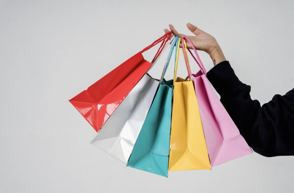 A person's hand holding a fan of colorful paper shopping bags in red, silver, teal, yellow, and pink colors against a plain light gray background.